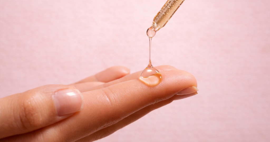 Glass dropper dispensing a drop of serum onto a fingertip against a soft pink background.