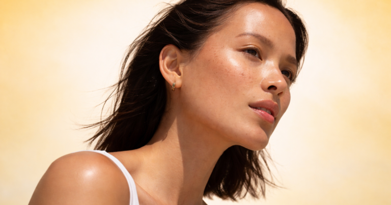Profile of a woman with glowing, dewy skin and natural freckles against a soft light yellow gradient background, with a clean skincare aesthetic.