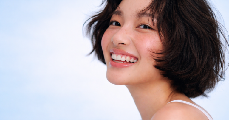 Smiling woman with glowing, dewy skin and short wavy hair against a soft light blue gradient background, with a clean skincare aesthetic.