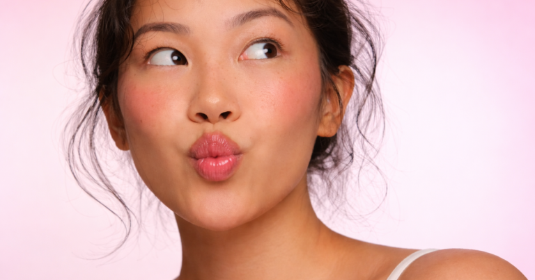 Close-up of a woman with natural glowing skin and freckles on a pink background.