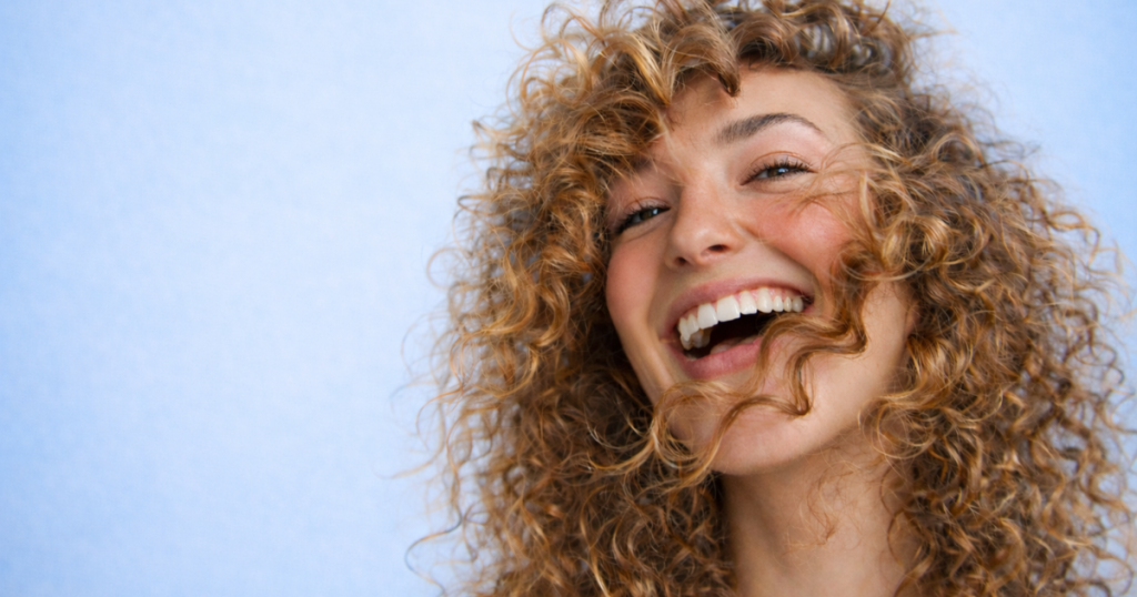 Smiling woman with natural curly hair and glowing skin against a soft light blue background.