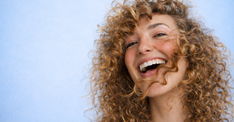 Smiling woman with natural curly hair and glowing skin against a soft light blue background.