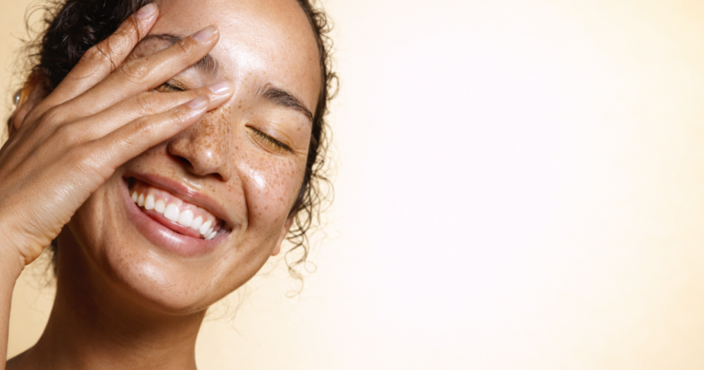 Smiling woman with radiant skin gently touching her face on a soft light yellow background.