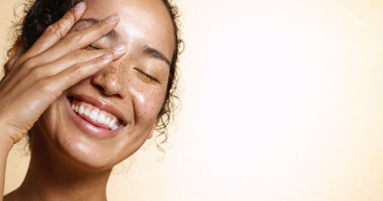 Smiling woman with radiant skin gently touching her face on a soft light yellow background.