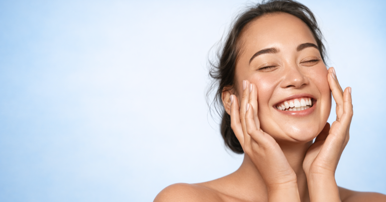 Smiling woman with radiant skin gently touching her face on a soft light blue background.