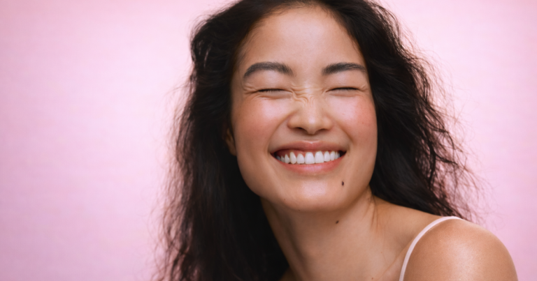 Smiling woman with long dark hair and closed eyes against a soft pink background.