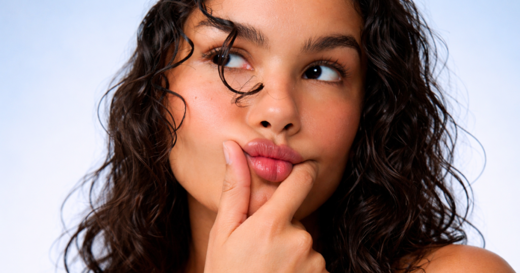 Woman with curly hair and natural skin on a soft blue background making a playful expression.