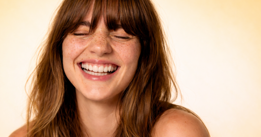 Woman with freckles smiling with eyes closed on a soft yellow background.