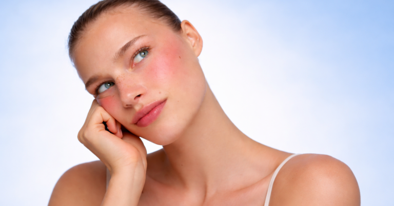 Portrait of a woman with glowing skin and a soft, natural smile, resting her face on her hand against a light blue gradient background.