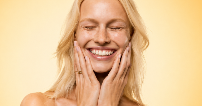 Smiling blonde woman with eyes closed, hands on her cheeks, against a soft warm yellow gradient background.