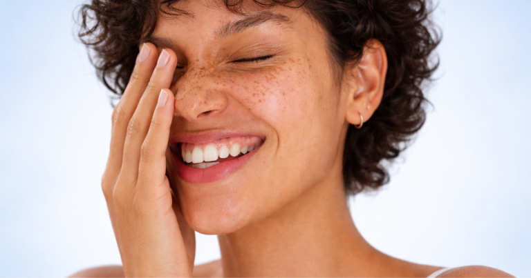 Close-up portrait of a smiling woman with freckles and short curly hair, eyes closed, gently touching her face, with soft natural lighting and a clean background.