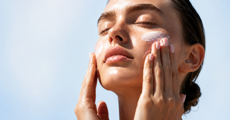 Woman with glowing skin and closed eyes applying sunscreen to her cheeks against a soft light blue sky background.