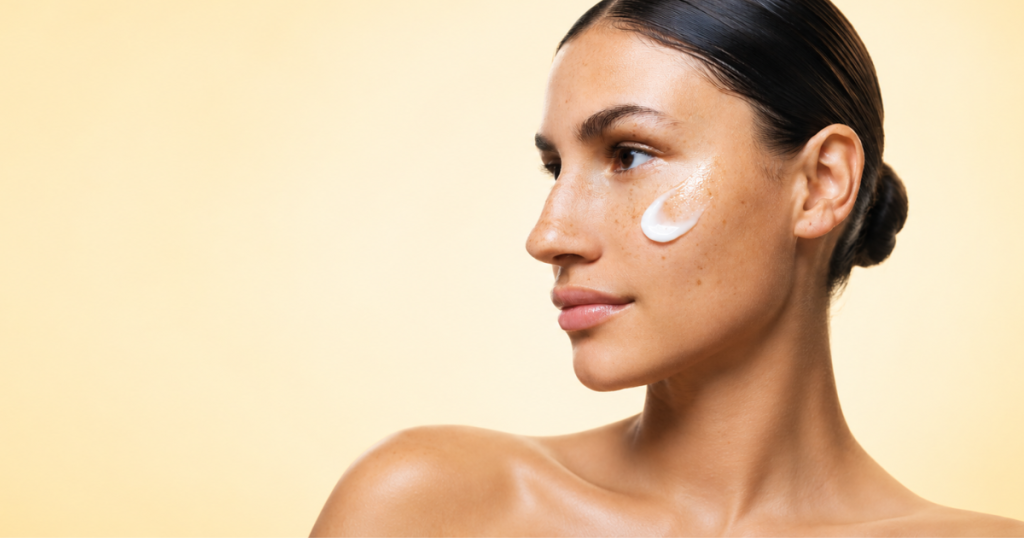 Close-up beauty portrait of a woman with glowing skin and freckles, wearing skincare cream on her cheek against a soft light yellow gradient background.