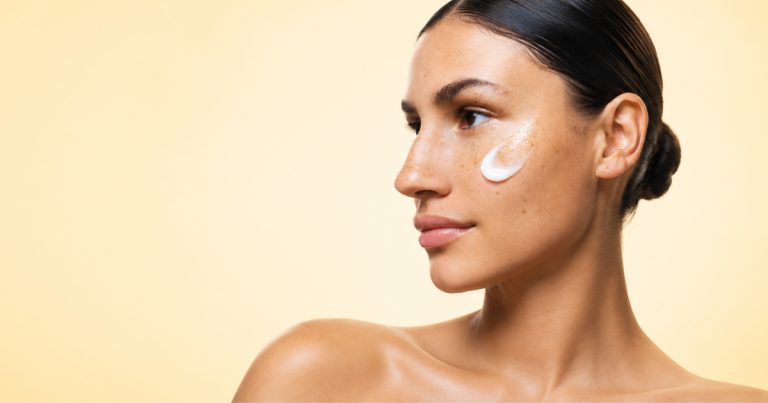 Close-up beauty portrait of a woman with glowing skin and freckles, wearing skincare cream on her cheek against a soft light yellow gradient background.