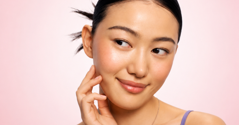 Portrait of a smiling woman with glowing skin and a natural makeup look against a soft blush pink gradient background that fades to a lighter center.