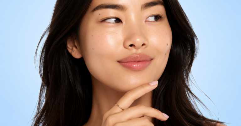 Portrait of a smiling woman with glowing skin and a natural makeup look against a soft blue gradient background that fades to a lighter center.