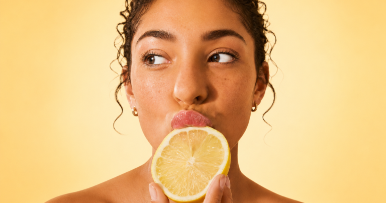 Close-up portrait of a woman holding a lemon slice to her lips against a soft yellow gradient background, with natural glowing skin and a playful expression.