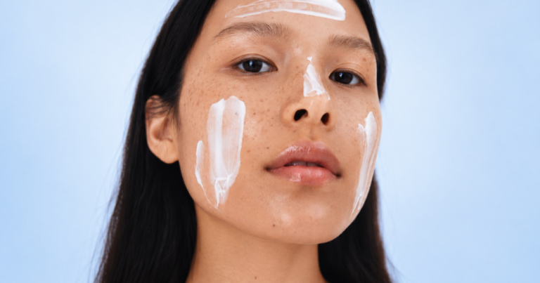 A woman with freckles and moisturizer applied to her face against a soft blue background.