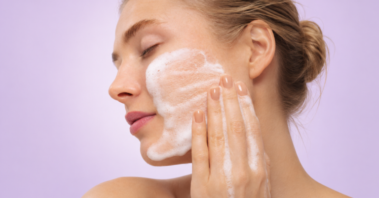 Woman washing her face with foaming cleanser against a soft lavender background.